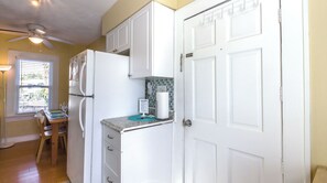 A compact kitchen with white cabinetry, a refrigerator, a countertop with a paper towel dispenser, and a window with blinds.