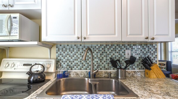 A kitchen with white cabinets, a stainless steel sink, a kettle, and a microwave.