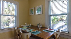 A dining area with a wooden table set for four, two framed artworks on the wall, and windows with blinds.