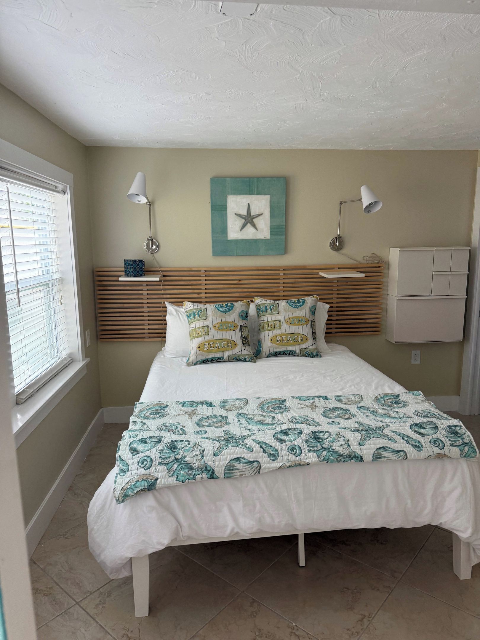 A bedroom with a bed, two bedside lamps, a wall-mounted shelf, a window with blinds, and a wall-mounted air conditioning unit.
