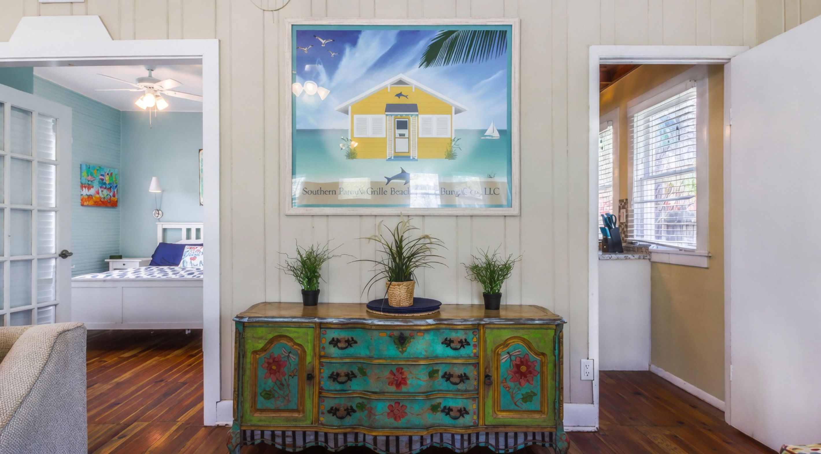 A room with a wooden floor, a colorful painted cabinet, a painting of a beach house, and a window with blinds.
