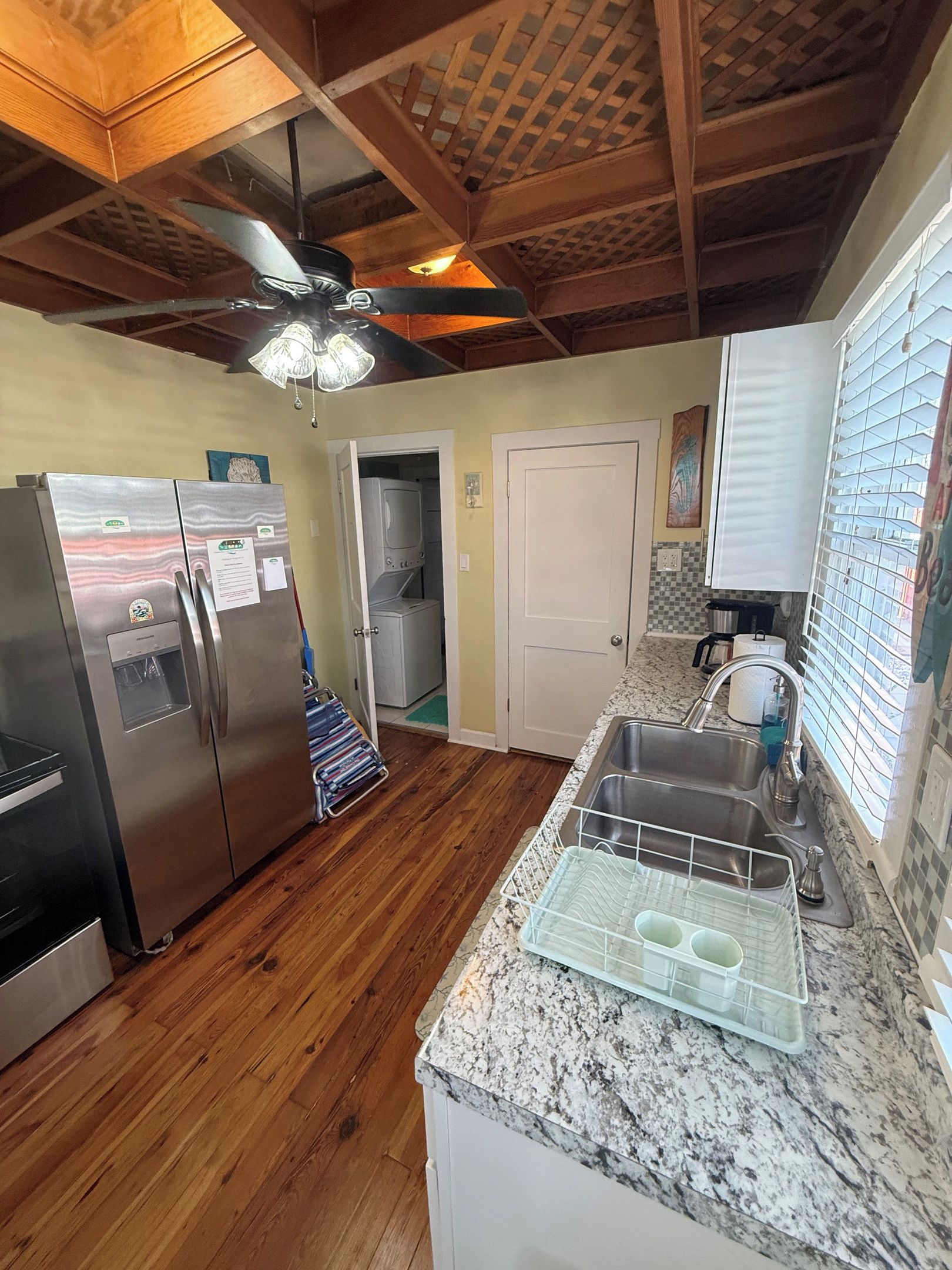 A kitchen with a stainless steel refrigerator, a built-in oven, a granite countertop, a sink with a dish rack, and a ceiling fan.