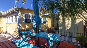 Une terrasse en bois avec une table de pique-nique et un parasol, entourée d’une maison jaune dotée d’une cour clôturée et de palmiers.