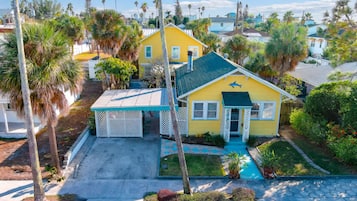 A yellow house with a covered porch, surrounded by palm trees and other vegetation.