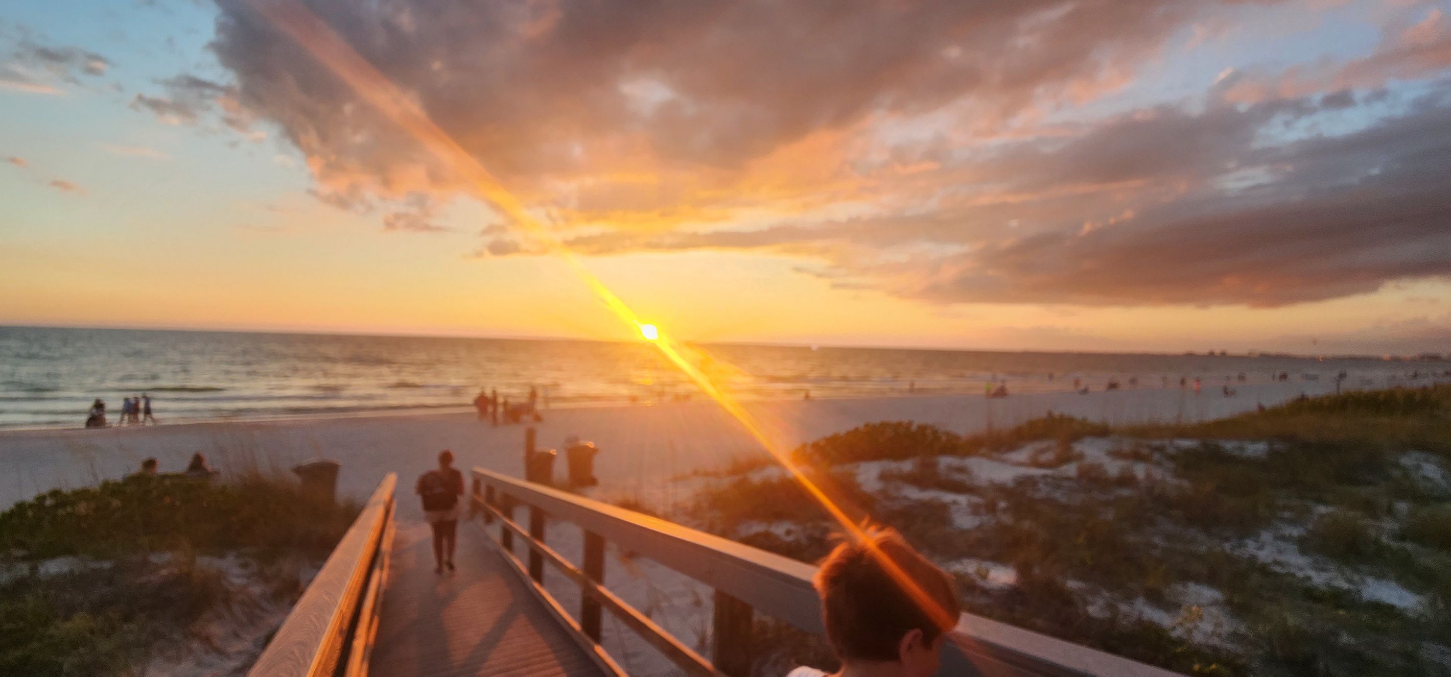 A sunset view from a boardwalk with people walking, a beach in the background, and a cloudy sky.