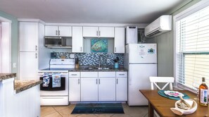 A compact kitchen with white cabinets, a microwave, stove, and a refrigerator. There's a window with blinds, a wooden table with a bottle, and a beach-themed mat.