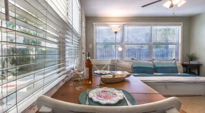 Un comedor con mesa de madera, sillas y banco. Hay ventanas con persianas, un ventilador de techo y una lámpara.