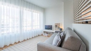 Ein modernes Wohnzimmer mit einem grauen Sofa, einem Flachbildfernseher und einem großen Fenster mit durchsichtigen Vorhängen.