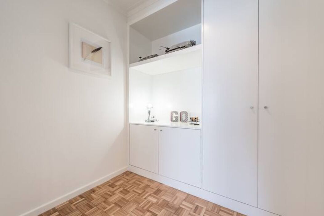 A room with a large wardrobe, a small shelf with decorative items, and a herringbone-patterned floor.