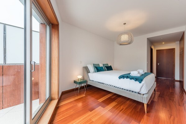 Une chambre à coucher moderne avec un grand lit, une table de chevet, un luminaire suspendu et une vue dégagée sur l’extérieur grâce à une grande fenêtre.
