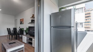 A modern living room with a flat-screen TV, a dining area with chairs, a coffee table, and a large stainless steel refrigerator.