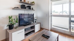 A modern living room with a flat-screen TV, wooden furniture, and a large window offering a city view.