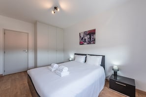 A neatly made bed with white linens and towels, a black nightstand with a lamp, a white wardrobe, and a cityscape painting on the wall. - GuestReady - Never Say Never (Matosinhos)