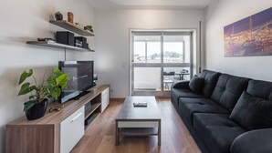 A modern living room with a flat-screen TV, a black sofa, a coffee table, and a balcony with a view of boats and a bridge.