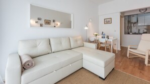 Een moderne woonkamer met een witte hoekbank, een eethoek met een kleine tafel en stoelen, en een keuken die zichtbaar is door een open doorgang.
