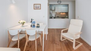 Een moderne, minimalistische eetruimte met een witte tafel, vier stoelen, een vaas met bloemen en een klok boven een keuken.