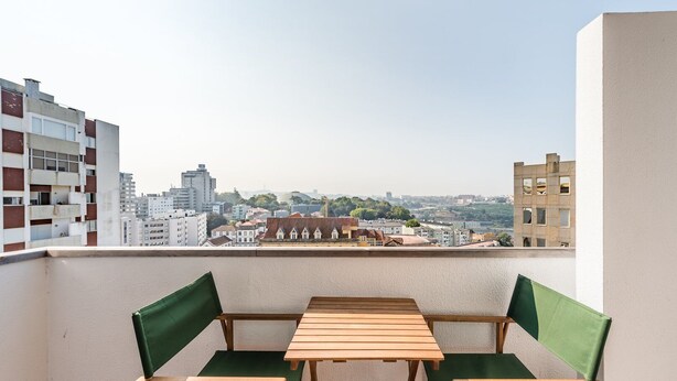 A balcony with a wooden table and green chairs, overlooking a cityscape with various buildings. - GuestReady - Urban escape with a view (Porto)