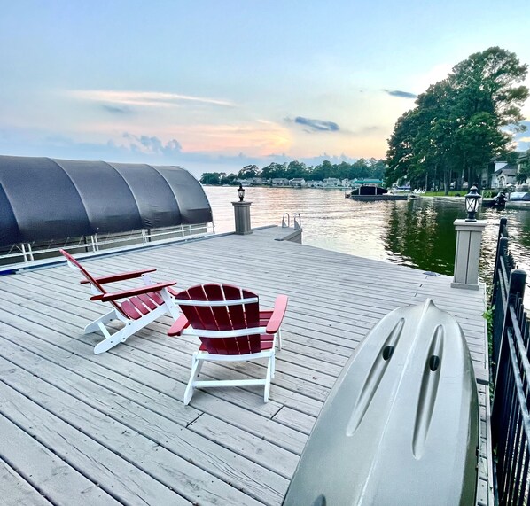 A wooden deck with Adirondack chairs and a grill, overlooking a calm body of water with a sunset in the background.