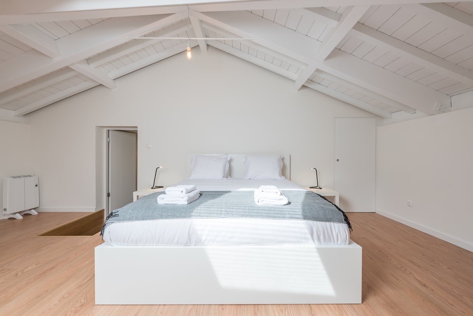 Un dormitorio moderno con techo inclinado, una cama grande con ropa blanca, una mesita de noche con lámpara y un radiador.