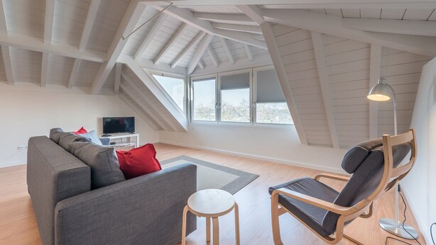 Una habitación moderna estilo loft con techo inclinado, ventanas amplias, un sofá gris, una silla de madera y una pequeña mesa.