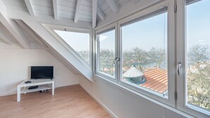 Habitación con techo inclinado, ventanas amplias, un mueble blanco para televisión y vistas a edificios y árboles.