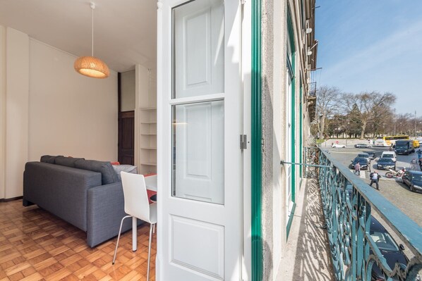 A balcony with a view of a busy street, a grey sofa, a white chair, and a hanging light fixture. - GuestReady - Clerigos-Virtudes Apartments (Porto)