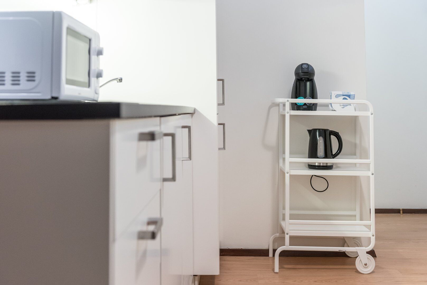 A kitchenette with a microwave, a coffee machine, and a white storage cart with shelves.