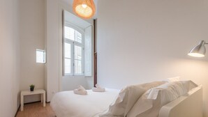 A minimalist bedroom with a large bed, a bedside table with a plant, a hanging light fixture, and a window with a view.