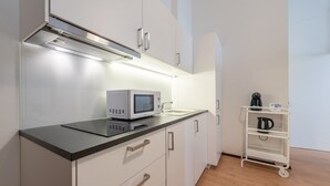 A modern kitchen with white cabinets, a black countertop, and a microwave. There is a white shelf unit with a kettle and coffee maker.