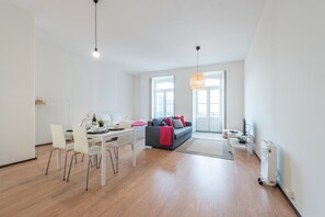 Uma sala de estar moderna com área de jantar, um sofá cinza com almofadas vermelhas e uma janela grande que proporciona iluminação natural.