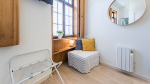 Une chambre à coucher de taille modeste, comprenant un lit, une table de chevet avec une lampe, une fenêtre avec des stores, un radiateur et un miroir mural.