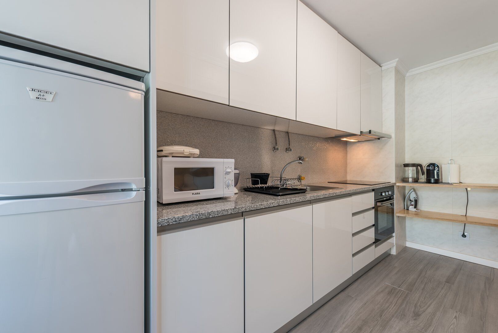 Een moderne keuken met witte kastjes, een ingebouwde magnetron, een spoelbak met kraan en een koffiezetapparaat op een plank.
