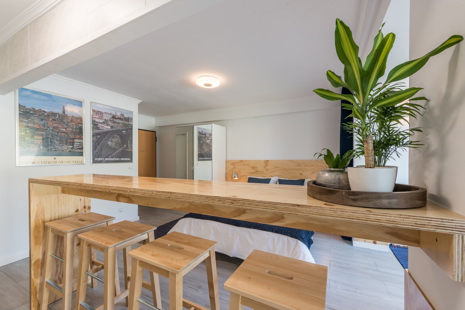 A modern, open-plan room with a wooden desk, bar stools, a large potted plant, and cityscape posters on the wall.