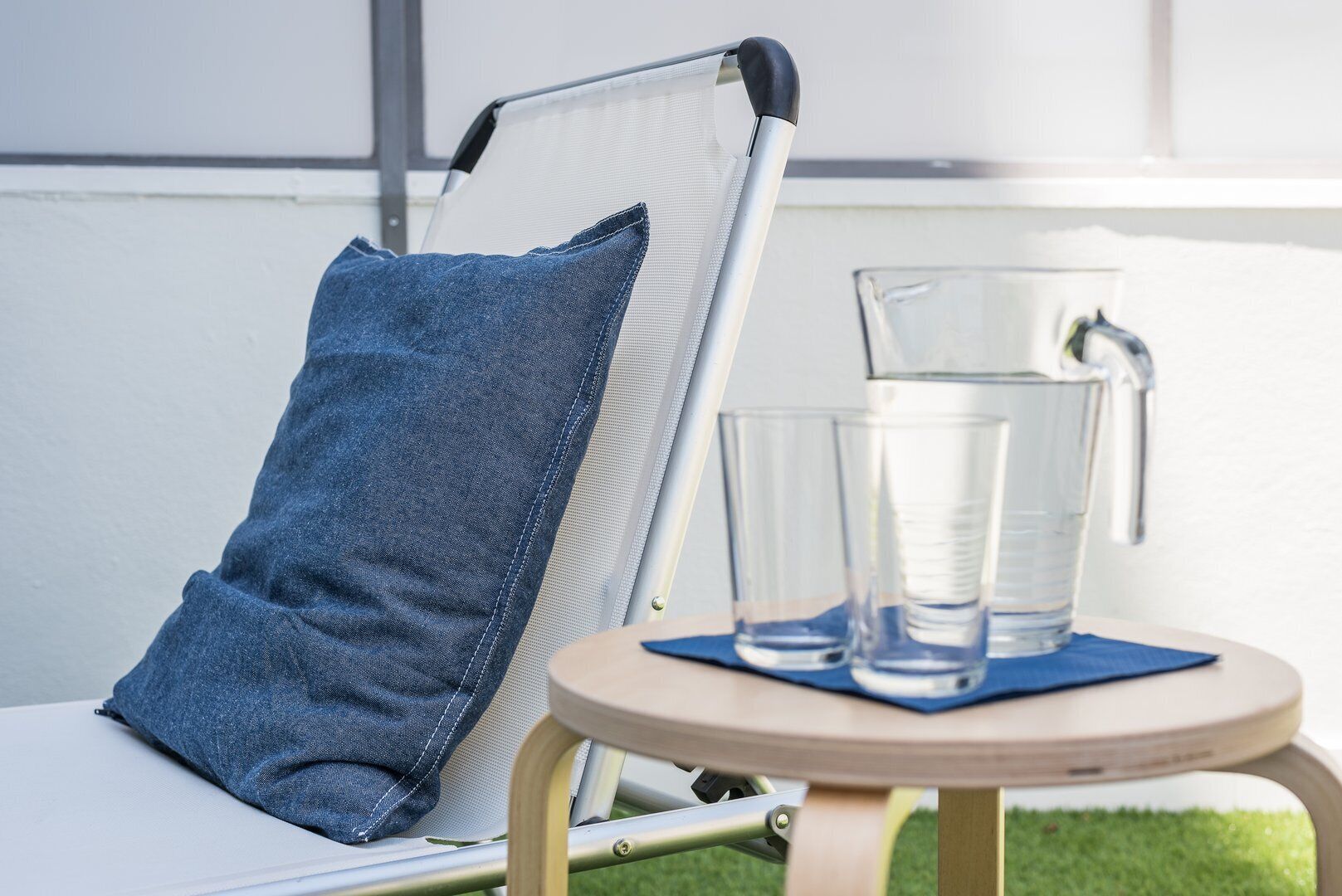 A chair with a denim pillow, a glass pitcher, and two glasses on a small round table.