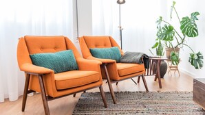 Two orange armchairs with teal cushions, a floor lamp, and a large green plant in a bright room with white curtains. - GuestReady - Chic and cozy getaway (Porto)