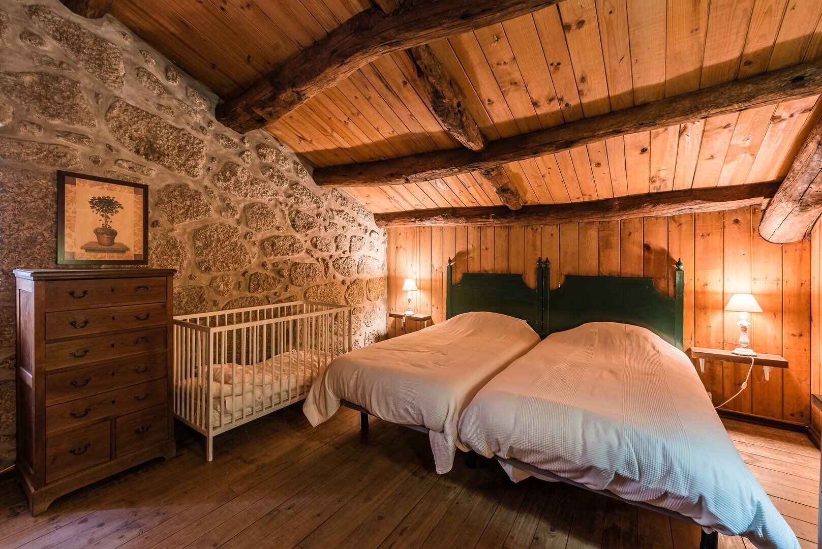 Un dormitorio rústico con pared de piedra, suelo de madera, una cuna, dos camas con cabeceros verdes y un aparador de madera.