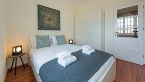 A neatly made bed with a blue blanket, two pillows, and folded towels on a grey headboard.