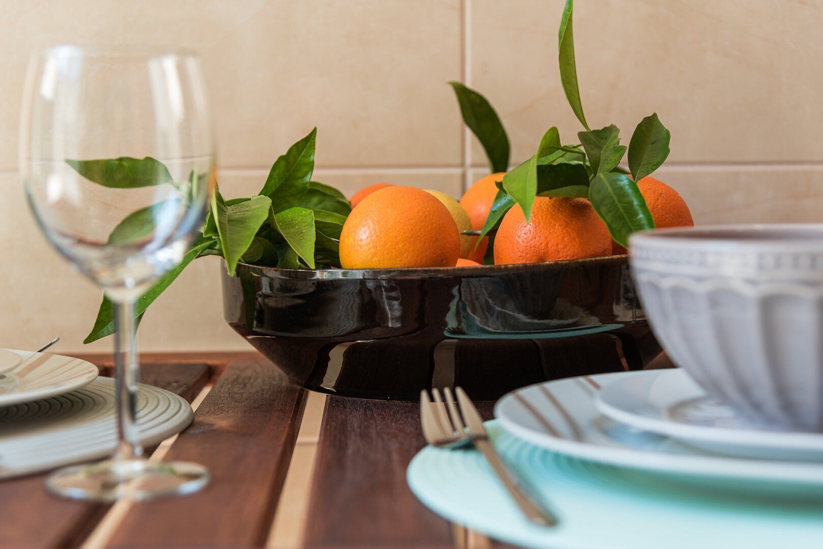 Une table à manger avec un bol noir contenant des oranges et des feuilles vertes, un verre à vin et un bol blanc.