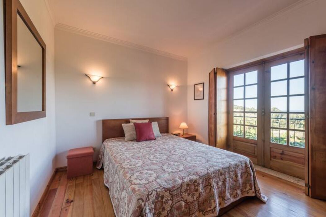 A bedroom with a large bed, wooden flooring, two bedside lamps, and a window with a view.