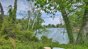 Un espacio de descanso junto al lago con dos sillas de metal y una mesa, rodeado de vegetación exuberante y árboles.