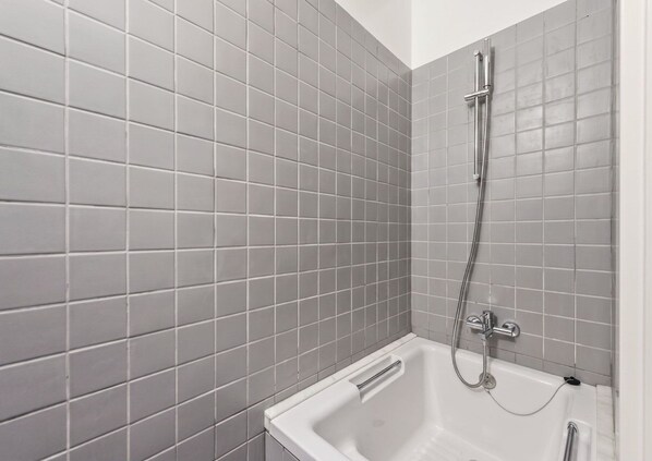 Ein Badezimmer mit grauen Fliesenwänden, einer weißen Badewanne und einer Dusche mit Duschbrause.