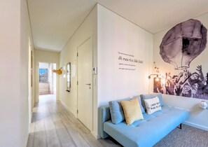 Un salon moderne avec un canapé bleu clair, une fresque murale et un couloir menant à une autre pièce.