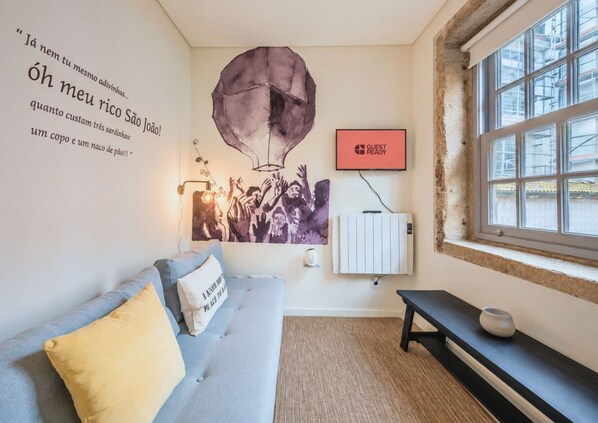A room with a grey sofa, a bench, a radiator, a window with scaffolding outside, and a wall with a poster and text. - GuestReady - Genuine Oporto Apts (Porto)