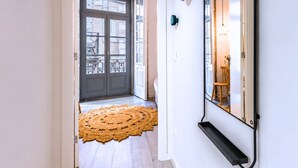 Un pasillo con una puerta oscura, un tapiz tejido, un estante en la pared y un espejo que refleja una habitación con una silla y una mesa.