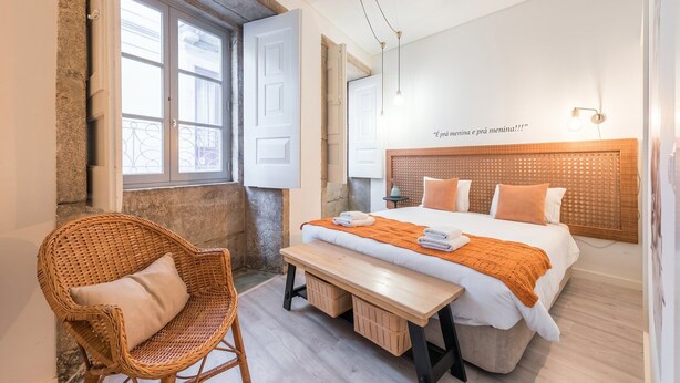 Een hotelkamer met een bed, een rieten stoel, een houten bankje en een raam met uitzicht.