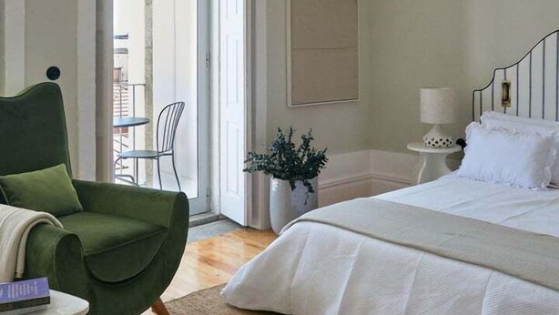 Une chambre à coucher avec un lit, un fauteuil vert, une petite table, une plante en pot et une vue sur l’extérieur.