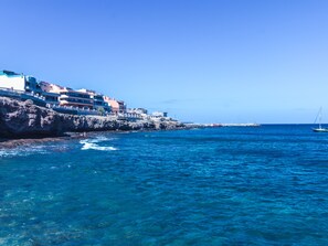 A coastal town with buildings along the rocky shore, clear blue sea, and a sailboat in the distance.