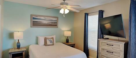 Habitación de hotel con cama, dos mesitas de noche con lámparas, ventilador de techo, televisor de pantalla plana, cómoda y un cuadro en la pared.