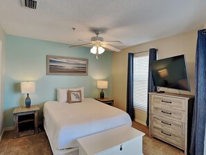 A hotel room with a bed, two bedside tables with lamps, a ceiling fan, a flat-screen TV, a dresser, and a painting on the wall.