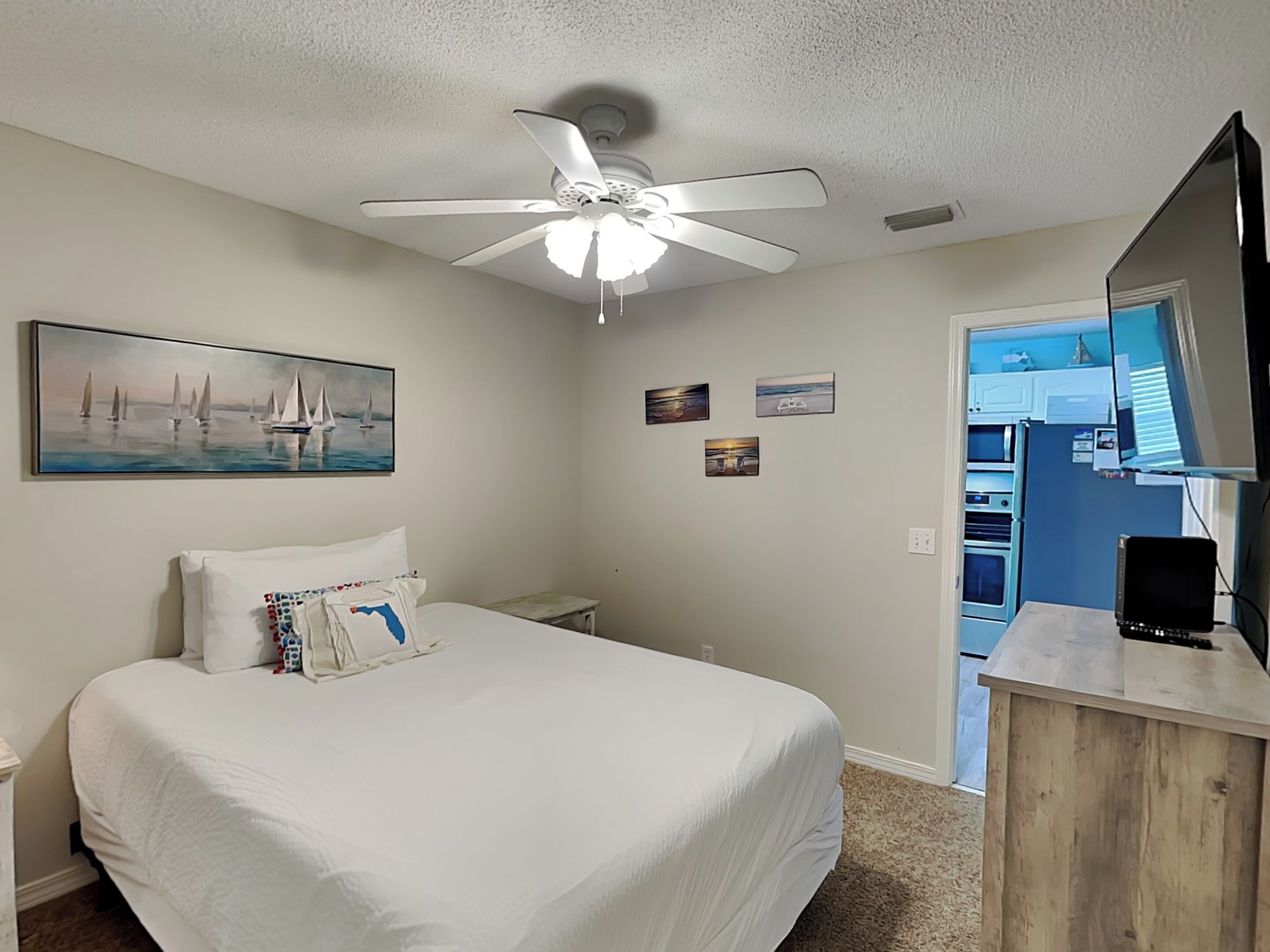 Una camera da letto con un letto, una ventola a soffitto, un quadro con dei velieri e vista su un angolo cottura.
